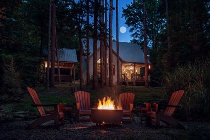 A night scene, fire glowing in fire pit, adirondak chairs surrounding, cabin in the background